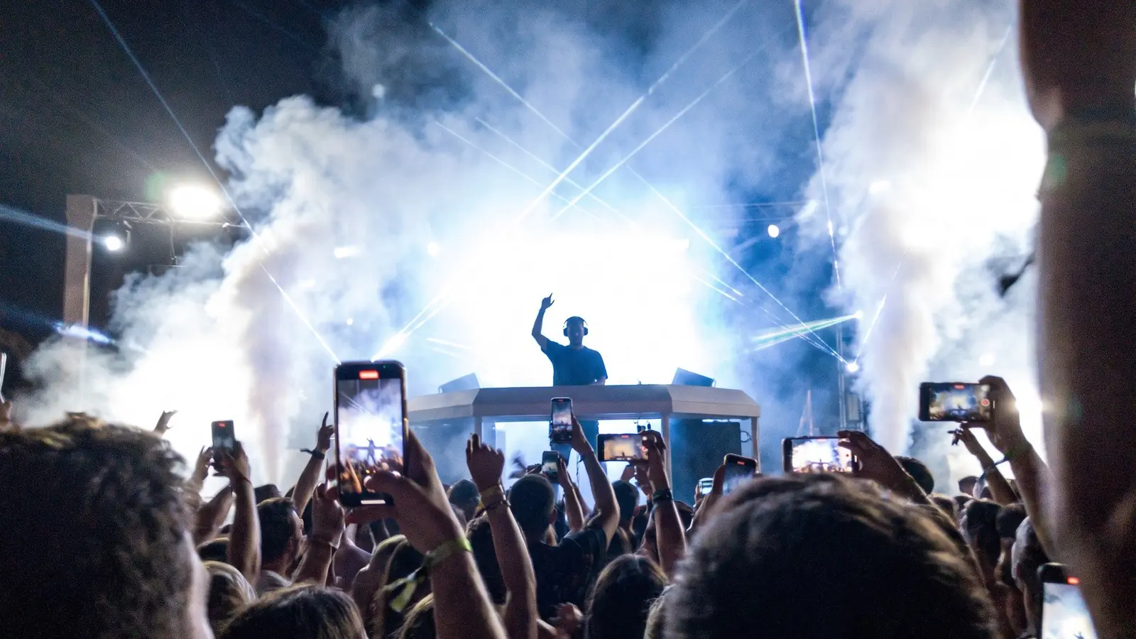 Cabina de DJ vista desde el público, con humo, luces azules y teléfonos en alto